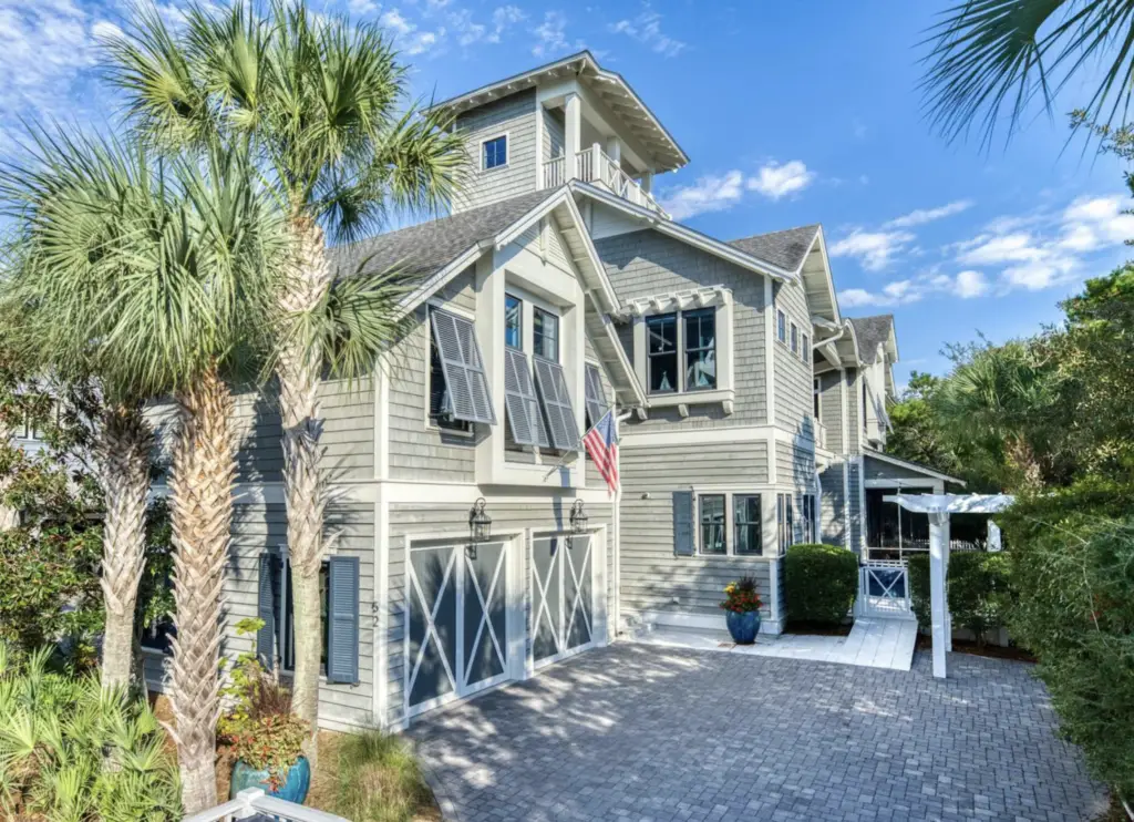 Watersound Beach Home - Living on 30A Florida Coastal gray beach house with white trim, palm trees, and a gated driveway under a blue sky with a flag.