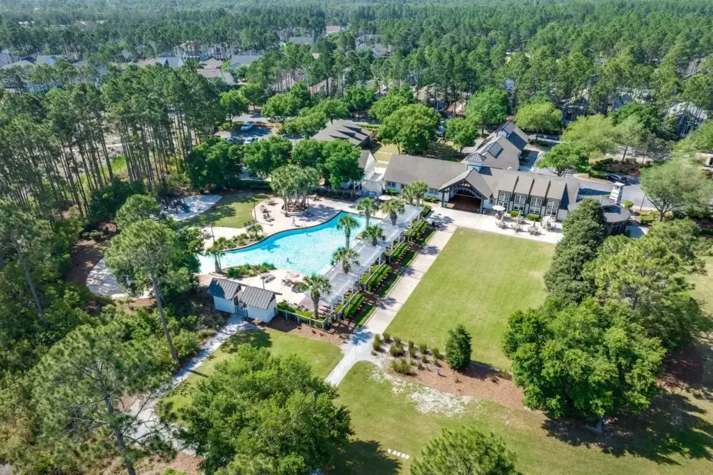 Aerial view of a resort-style pool area with palm trees, lounge chairs, and a shaded pergola beside a clubhouse, surrounded by pine woods.
