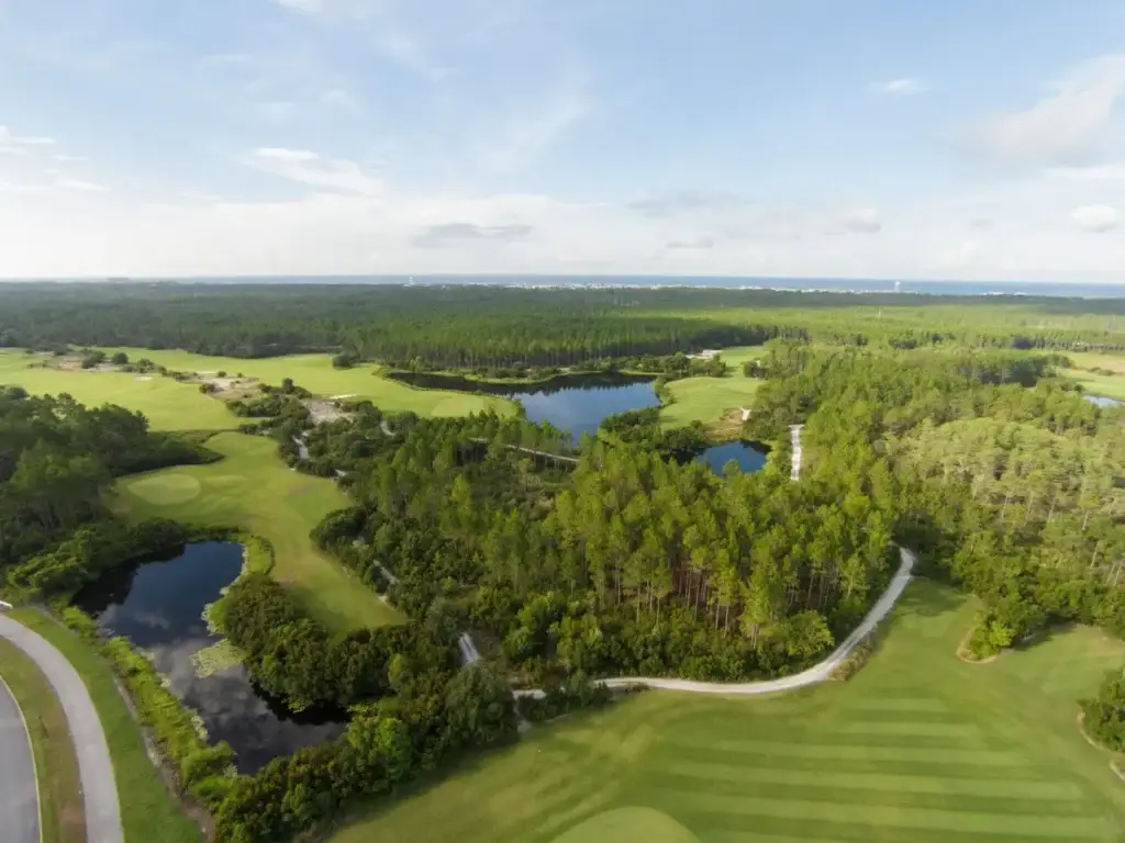 Aerial view of Watersound Origins Golf Course with ponds, winding cart paths, and surrounding trees reaching toward the ocean.